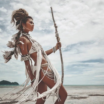 Vrouw in witte boheemse strandkledij met veren en houten staf op een rustige kust onder bewolkte hemel
